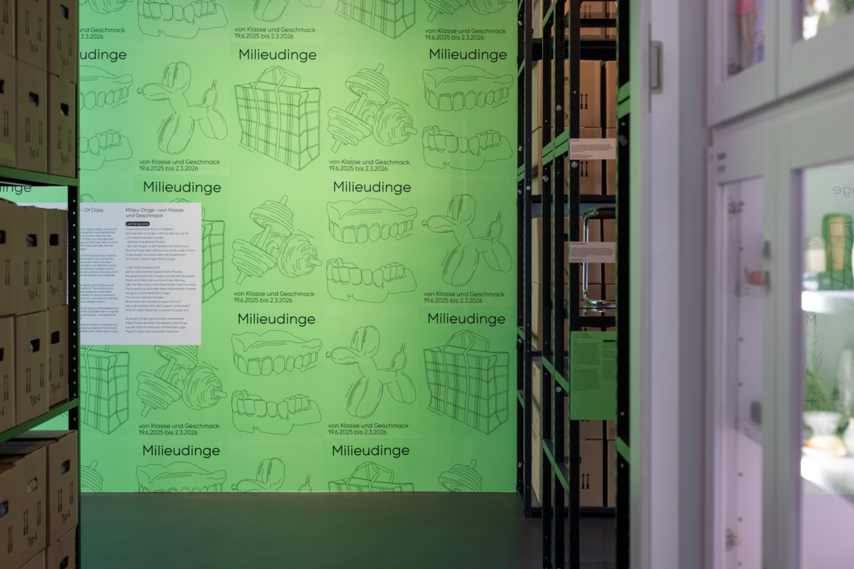 Room image with cabinets with showcases and shelves with packing boxes on both sides. In the background is a wall covered in bright green posters with the key visuals: dentures, dog balloon, chequered bag and dumbbells.