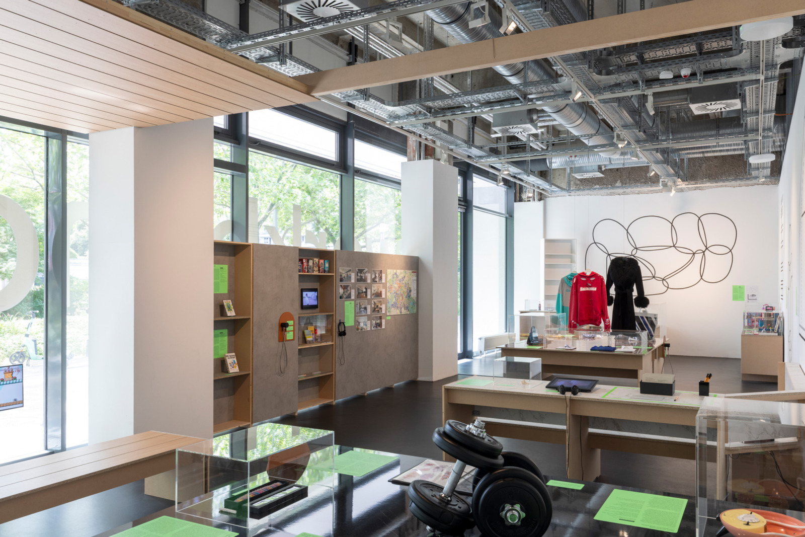Exhibition room with various objects and furniture: dumbbells, long black vintage coat, press-on nails, tattoo sheets in a folder.