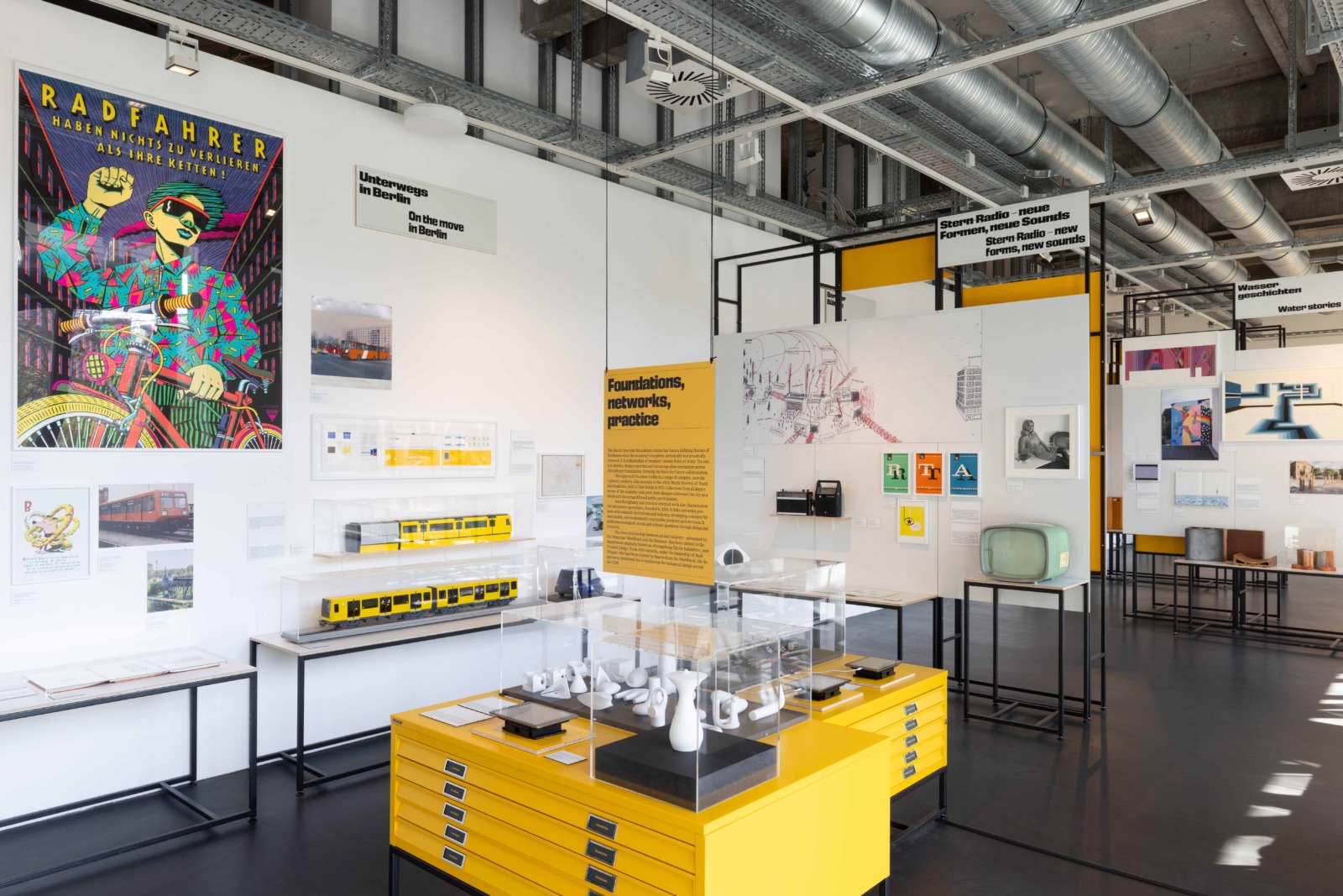 An exhibition area on the topic of mobility in Berlin, featuring models of subway trains in display cases, a colorful “Cyclist” poster, and various design objects