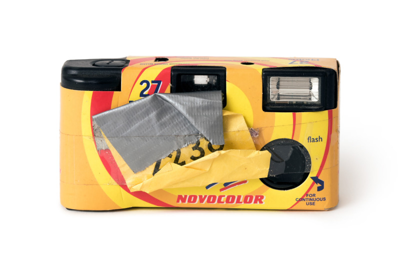 A yellow-and-red Novocolor disposable camera against a white background, the front of which is partially covered with silver duct tape and an attached clothing tag.