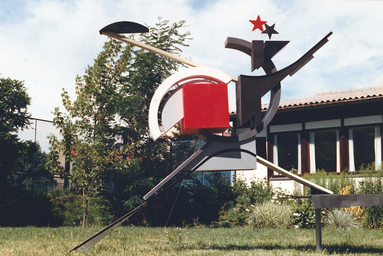 An avant-garde, constructivist sculpture featuring abstract geometric shapes—such as a red square and two stars at the top—stands on a lawn outdoors.